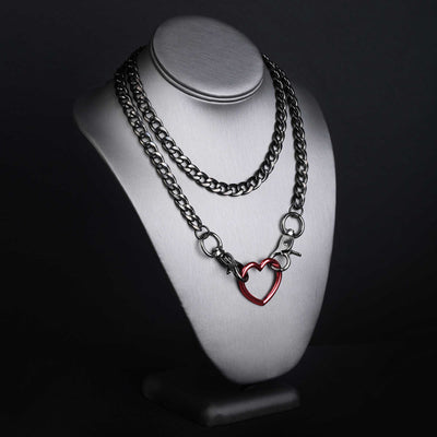 A double-layered chain necklace displayed on a white neck mannequin against a dark background. The top layer features a silver-toned curb chain, and the bottom layer features a silver-toned cable chain with a red heart-shaped pendant.