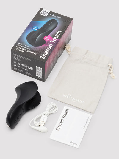 WeVibe Shared Touch dual-person intimate massager set with black handheld device, white USB charging cable, beige drawstring pouch, and printed box and booklet on a light gray background.