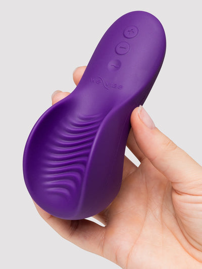 A hand holding a purple personal massager shaped like an insole, with three control buttons on the top surface, against a light gray background.